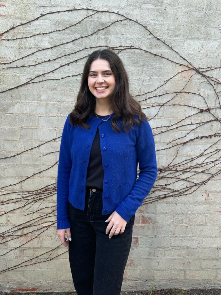 A woman with long dark hair, wearing a blue cardigan and black pants, stands smiling in front of a vine-covered, light-colored brick wall. This could be none other than Caoimhe Lyons.