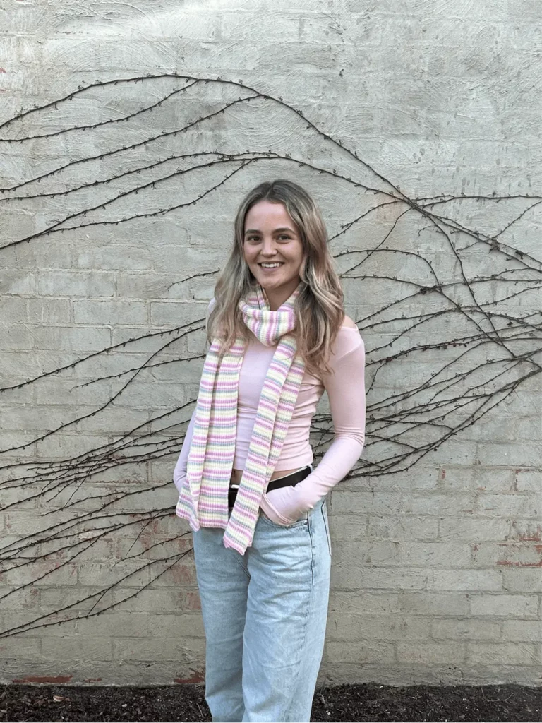 Chelsea, A smiling woman with long hair stands in front of a brick wall with climbing vines, wearing a pink long-sleeve shirt, light blue jeans, and a pink striped scarf.