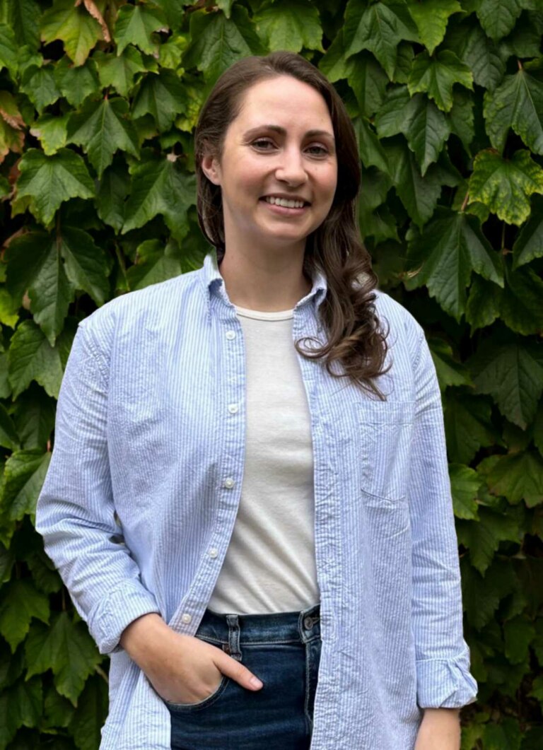 A woman in a blue striped shirt and jeans smiling in front of a leafy green background, photographed by Annelies Kersemakers.
