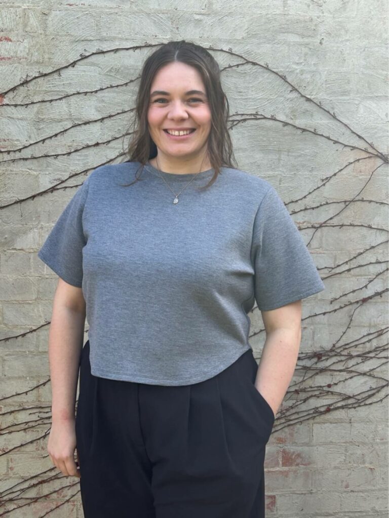 Jodie Thom stands against a brick wall with bare vine branches, wearing a gray t-shirt and black pants, smiling at the camera.