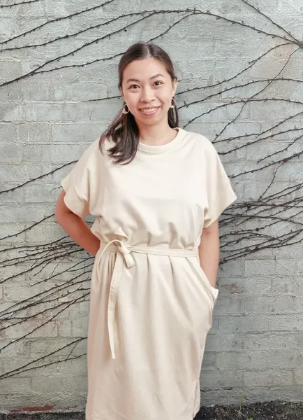 Sheila Mendoza stands smiling against a brick wall adorned with vine patterns, wearing a light-colored dress cinched with a belt.