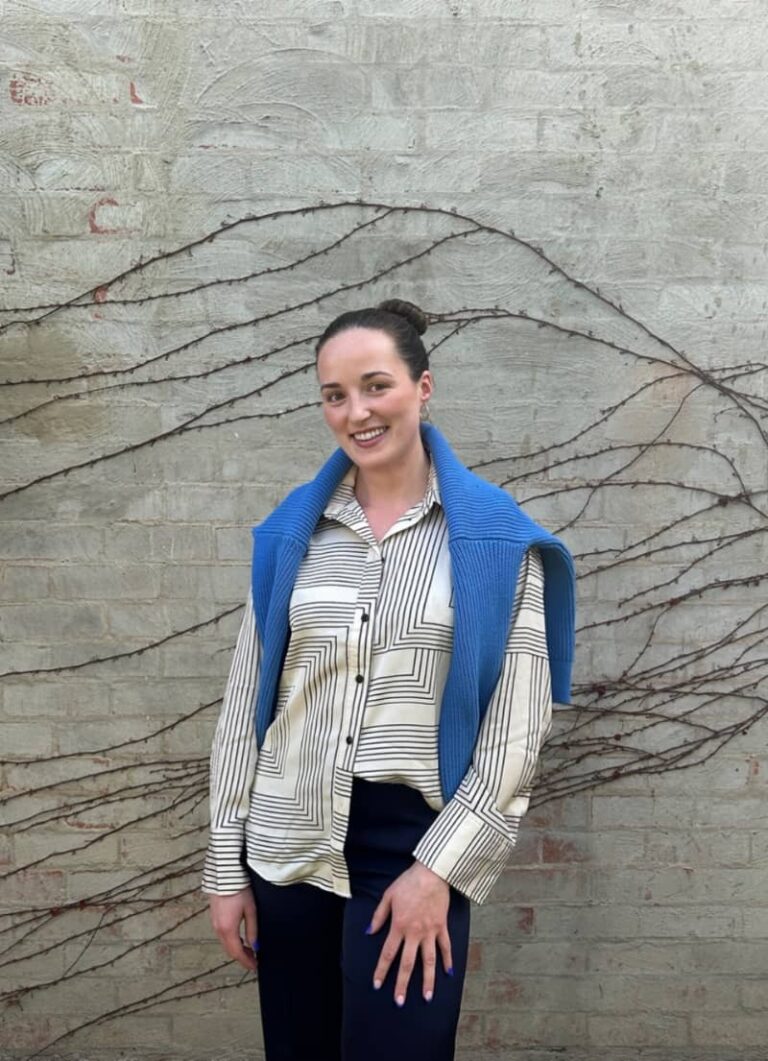 Sinead Scott stands in front of a textured wall with vine-like details, wearing a patterned shirt and a blue sweater draped over her shoulders, smiling at the camera.