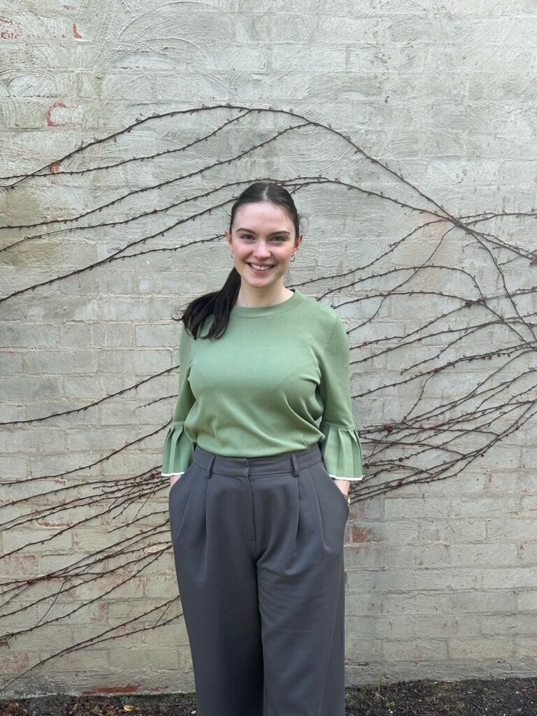 A person with long hair in a ponytail, wearing a green Maccora top and grey pants, stands in front of a brick wall with vine patterns.