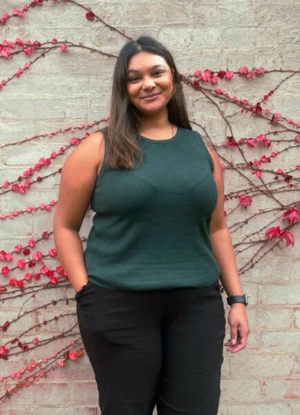 A woman wearing a dark green sleeveless shirt and black pants stands in front of a brick wall adorned with red vines, reminiscent of an Aashia Deo advert.