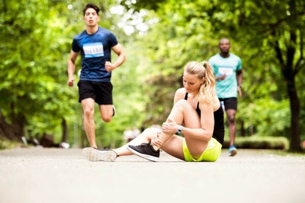 A woman is sitting on the ground worrying about her sprained ankle while a man is jogging.