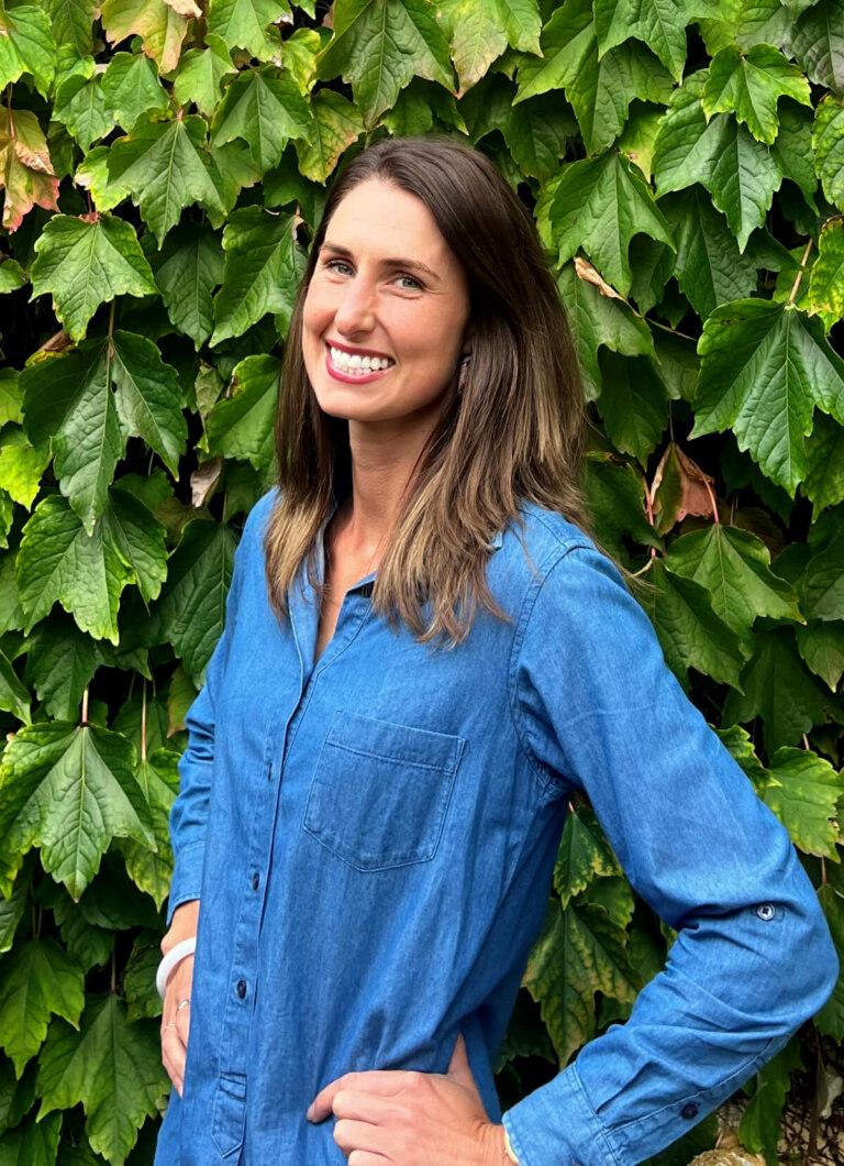 A woman in a blue shirt standing in front of a bush - Riandri Roux.